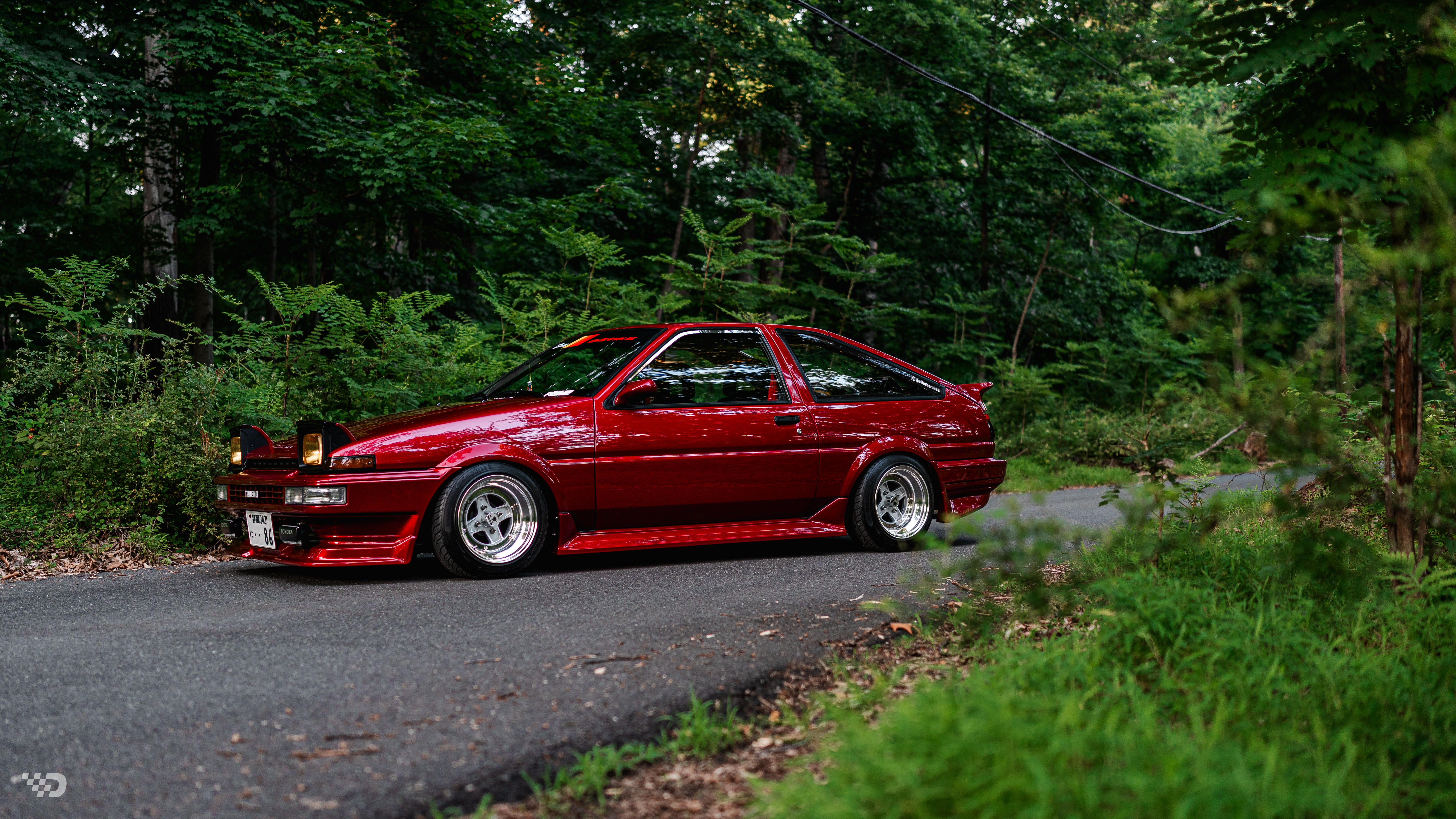 Red 1985 Toyota Corolla GTS (AE86) parked on a wooded road with pop-up headlights raised.