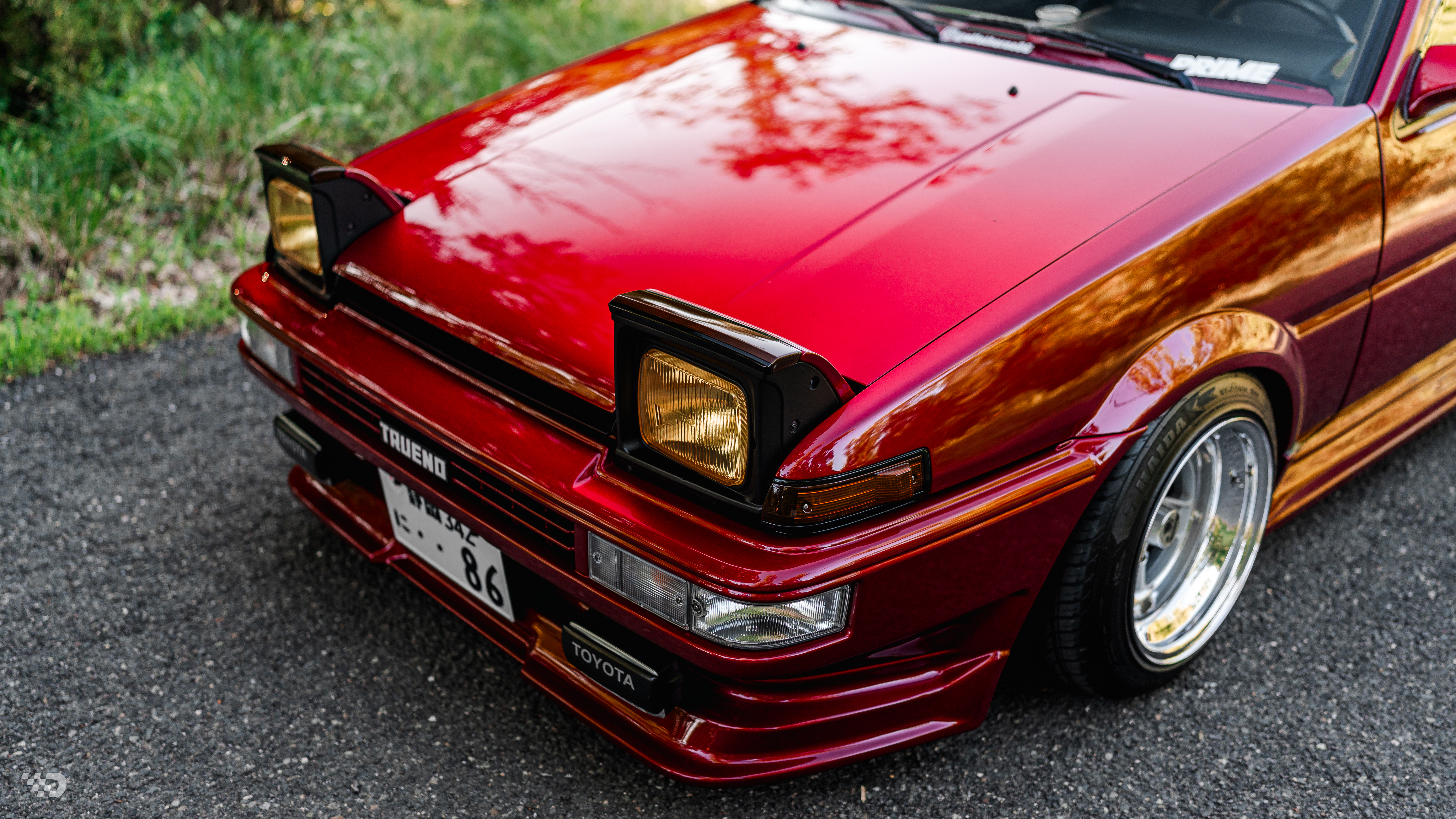 Front three-quarter detail of red AE86 Trueno with pop-up headlights raised and polished lip wheels.
