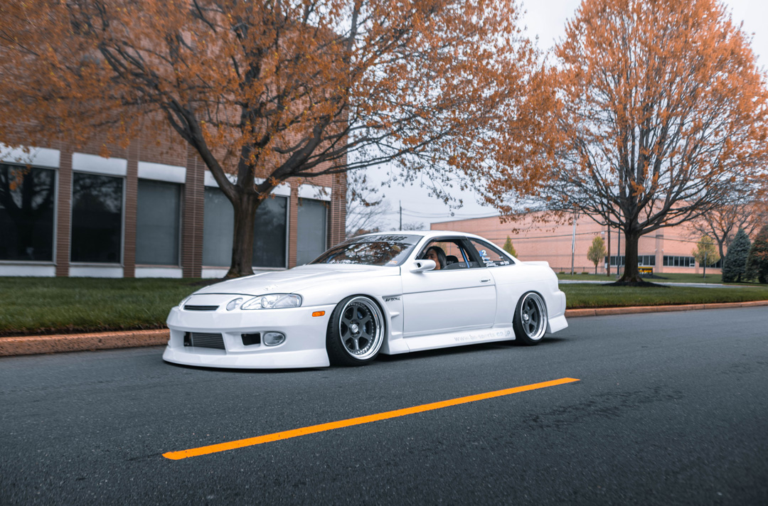 Aum Sung’s 1992 Lexus SC400 featuring BN Sports widebody styling, lowered stance, and Work Meister wheels driving through an urban street setting.