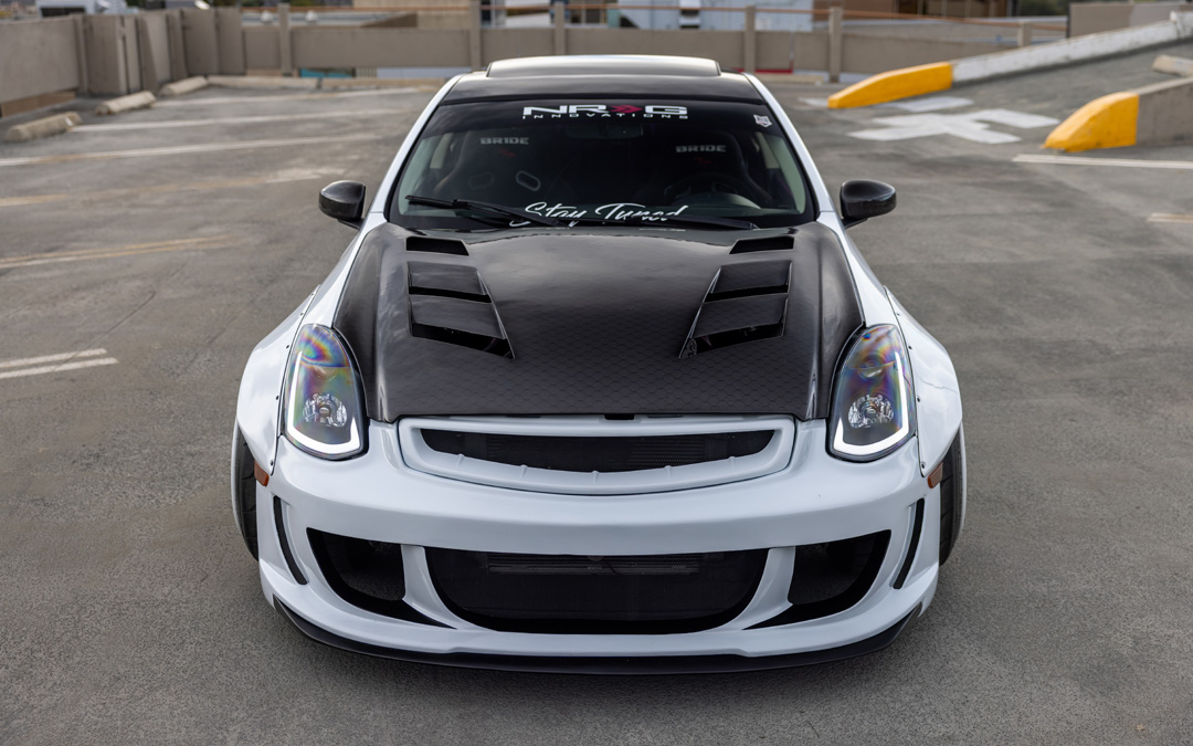 Front view of Jordyn Brazeau’s white widebody 2006 Infiniti G35 showing the carbon hood, custom headlights, front bumper, and wide fender fitment.
