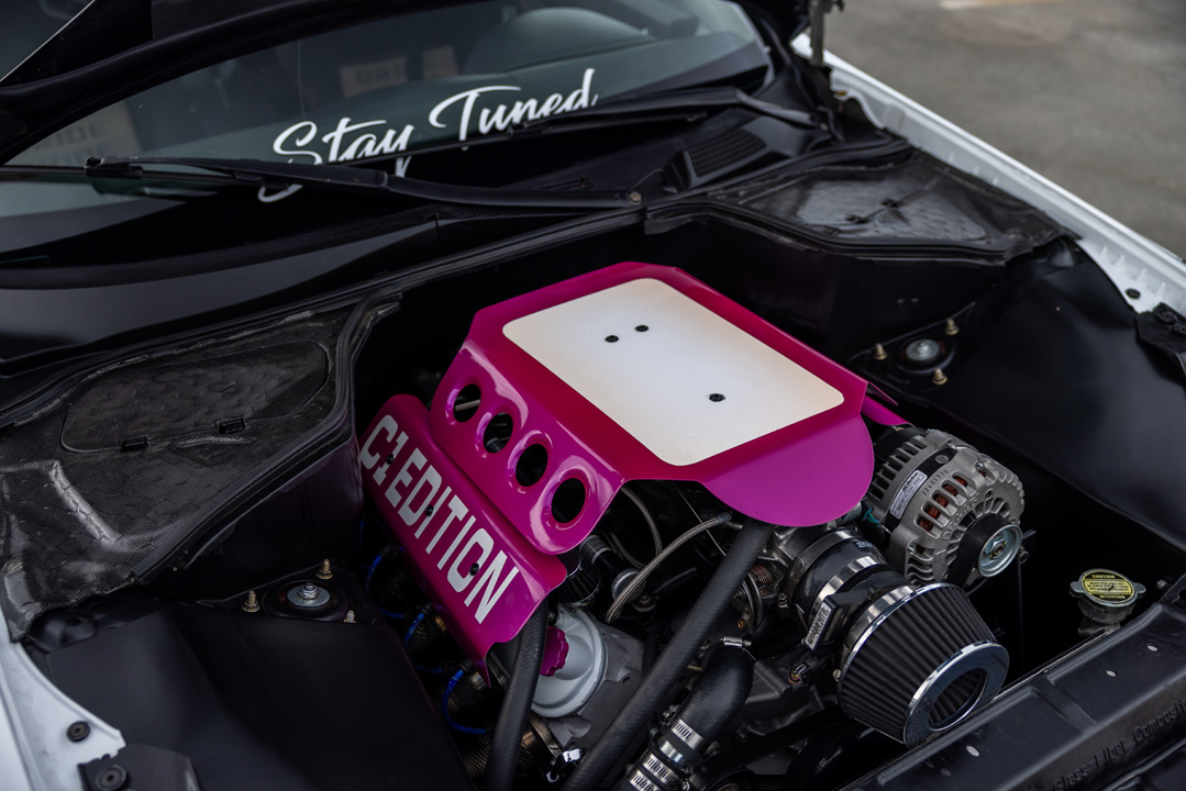 Engine bay detail of Jordyn Brazeau’s 2006 Infiniti G35 showing the LS swap, pink Chevrolet engine cover, custom shaved bay, and Stay Tuned windshield banner.