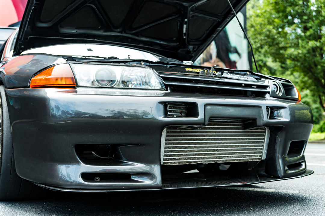 Front bumper detail of Cee Vann’s gray 1990 Nissan Skyline GT-R showing the exposed front-mount intercooler, carbon splitter, and open hood.