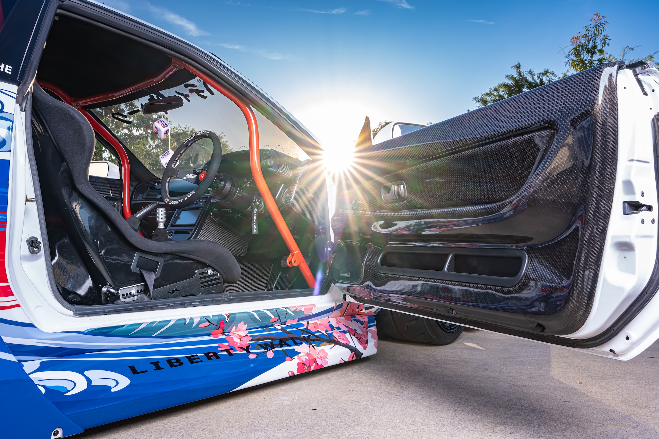 Interior detail of Osmond Seangsuwan’s 1990 Nissan Skyline GT-R showing the Garage Active carbon door panel, Sparco racing seat, red Cusco roll cage, Liberty Walk livery, and custom cockpit.