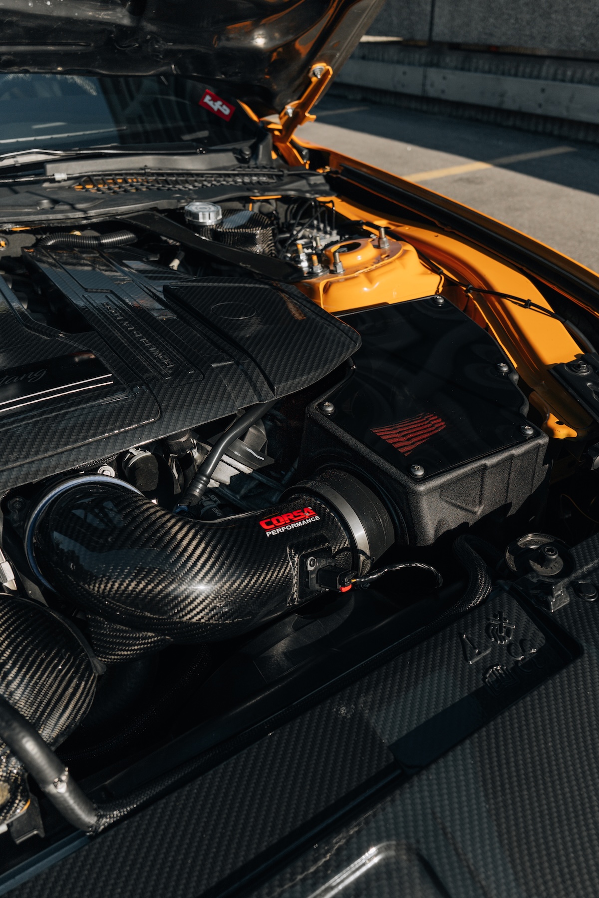 Close-up engine bay detail of Mussi Hussain’s 2022 Ford Mustang GT showing the Corsa carbon-fiber cold-air intake, carbon-fiber engine dress-up pieces, and Cyber Orange engine bay accents.