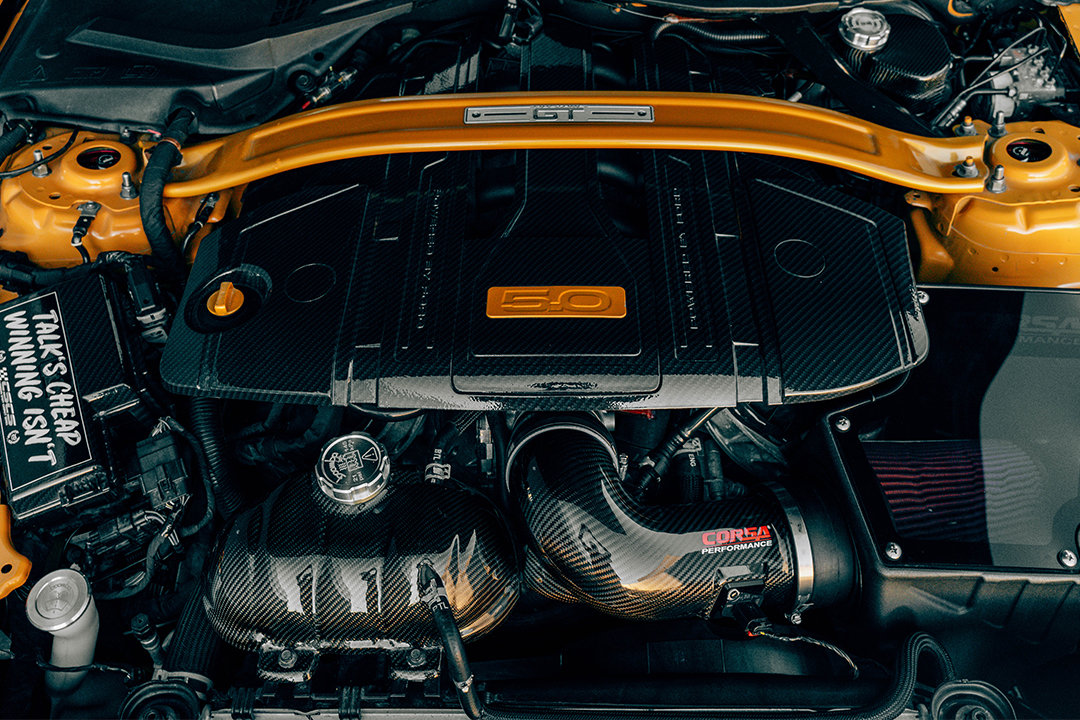 Engine bay detail of Mussi Hussain’s 2022 Ford Mustang GT showing the Coyote 5.0 engine, carbon-fiber dress-up pieces, Corsa carbon-fiber intake, and Cyber Orange strut bar.