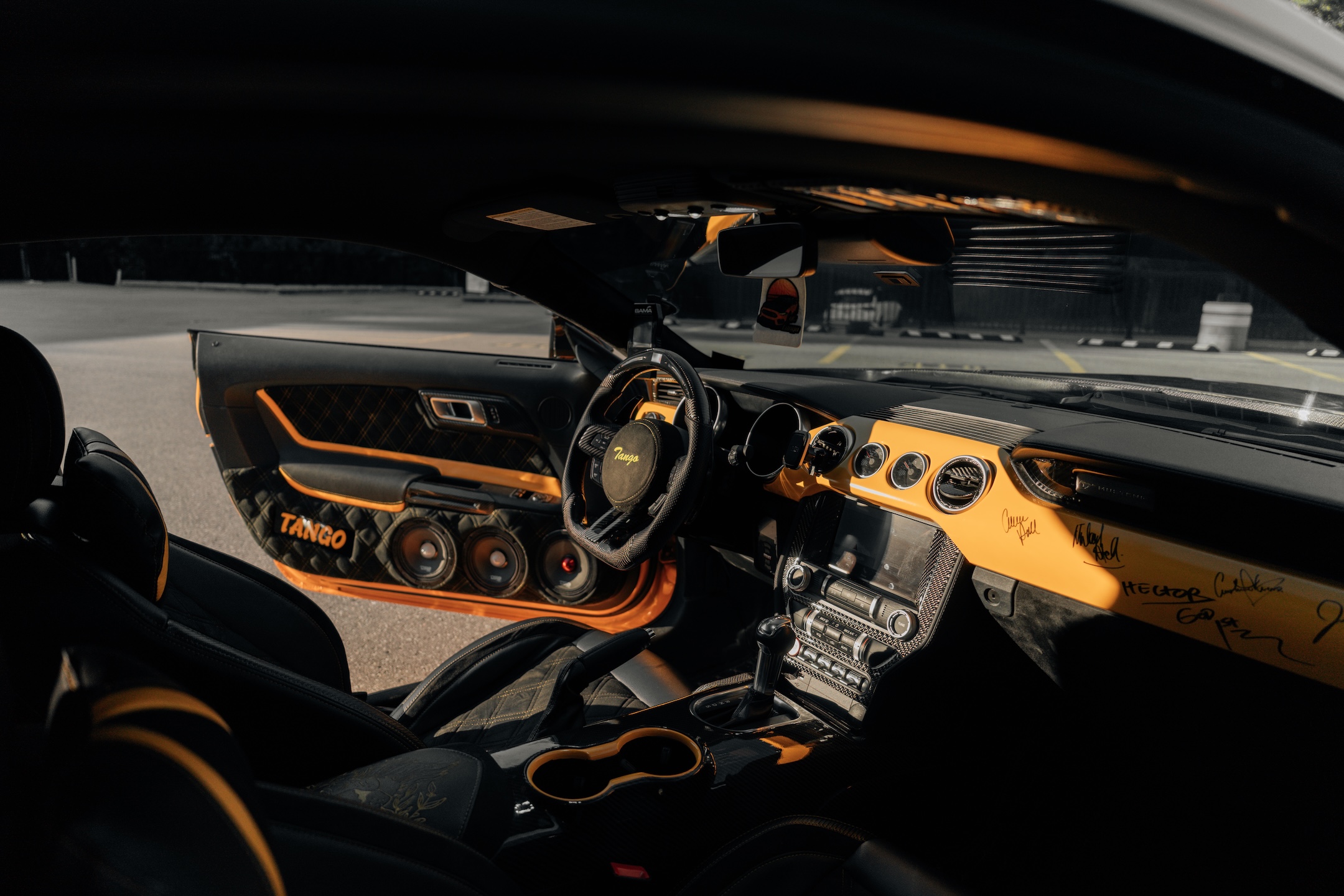 Interior detail of Mussi Hussain’s 2022 Ford Mustang GT showing the custom orange and black cabin, carbon steering wheel, Cyber Orange trim, custom door panel speaker pods, and diamond-stitched upholstery.
