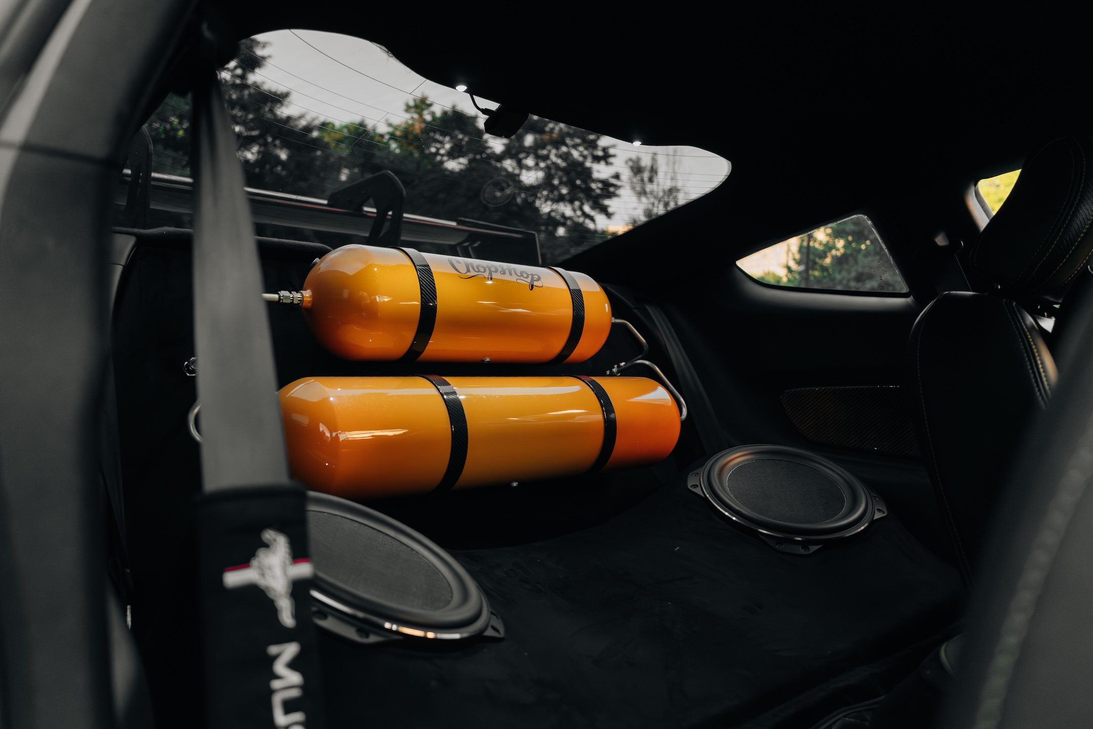 Rear-seat delete detail of Mussi Hussain’s 2022 Ford Mustang GT showing the ChopShop air suspension tank setup, custom audio layout, orange air tanks, and black upholstered rear platform.
