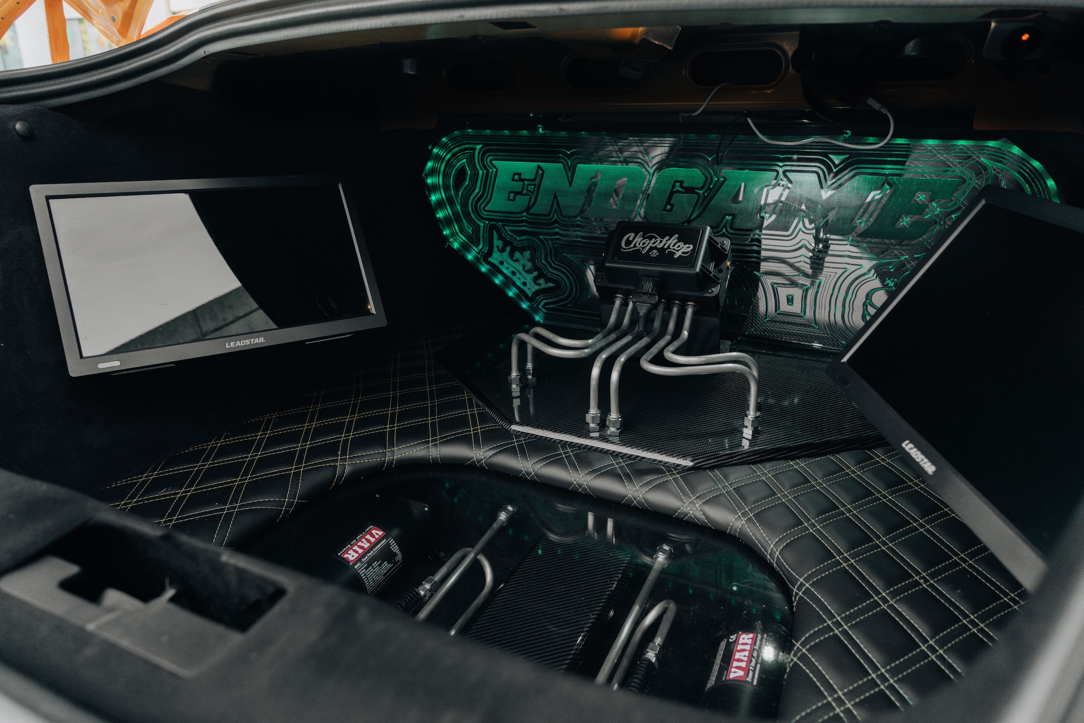 Custom trunk setup in Mussi Hussain’s 2022 Ford Mustang GT showing the ChopShop air suspension hardline setup, carbon-fiber platform, integrated screen, EndGame display panel, and quilted upholstery.