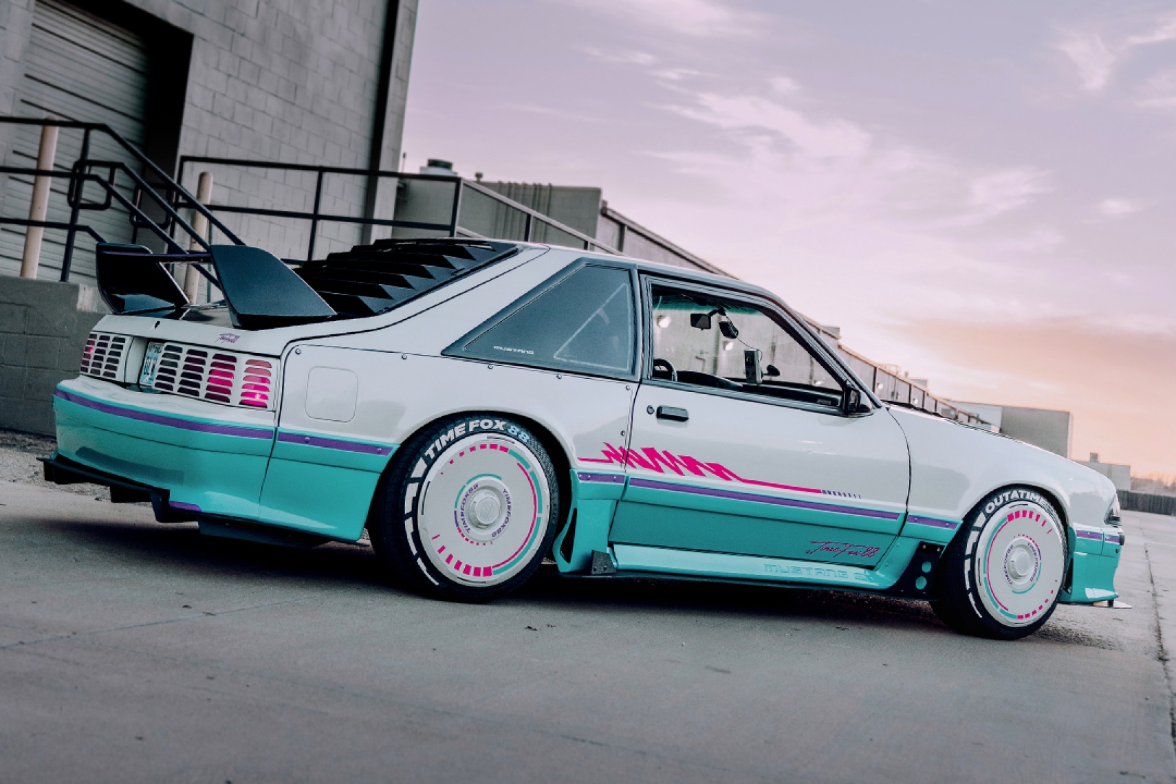 Rear three-quarter view of Regan McLaughlin’s 1988 Ford Mustang GT TimeFox showing the custom multi-tone paint, wide fiberglass fenders, Mustang rear window vents, Evo-style rear wing, HaloBLK wheel covers, and retro Foxbody styling.