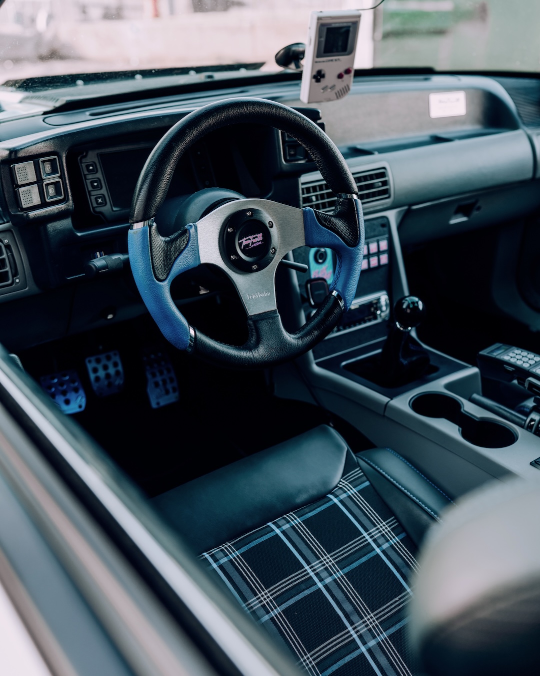 Interior detail of Regan McLaughlin’s 1988 Ford Mustang GT TimeFox showing the blue plaid seats, NRG steering wheel, Haltech keypad controller, Game Boy digital speedometer, and retro-inspired Foxbody cabin.