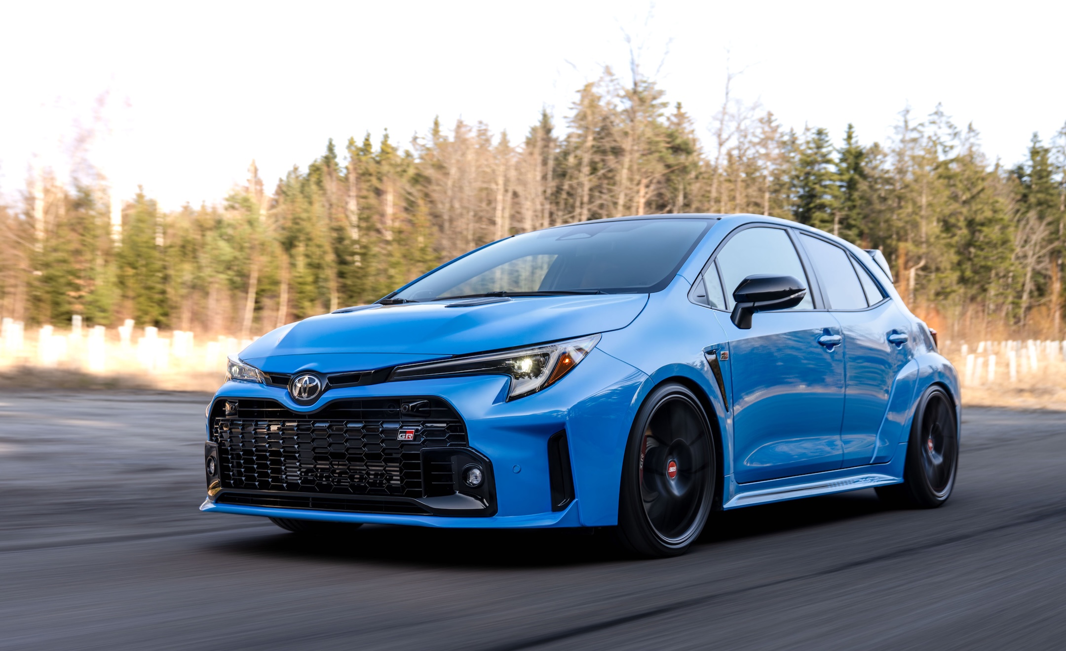 Blue Toyota GR Corolla equipped with ST XTA plus 3 coilover suspension driving on a road from a front three-quarter angle.