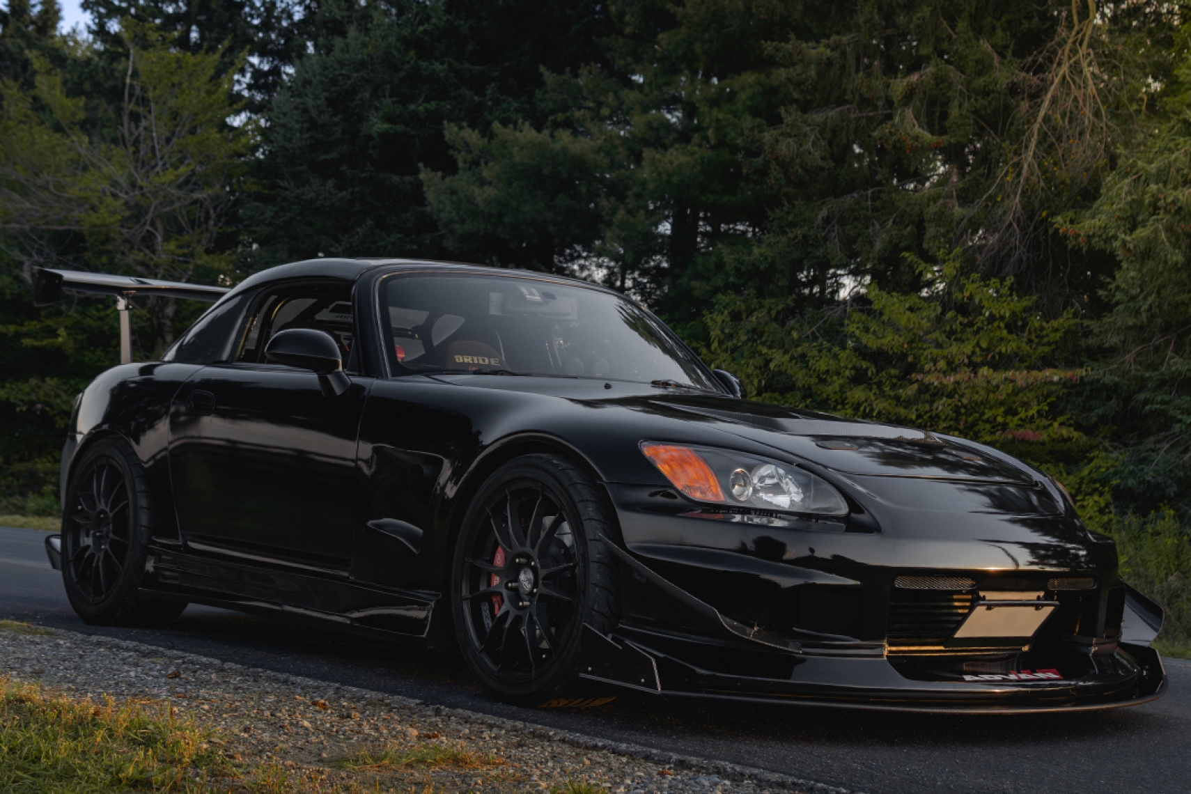 Front three-quarter hero view of Thomas Wu’s black 2002 Honda S2000 showing the turbo track build, Voltex-style aero, front splitter, hardtop, rear wing, OZ Racing wheels, and wooded road setting.